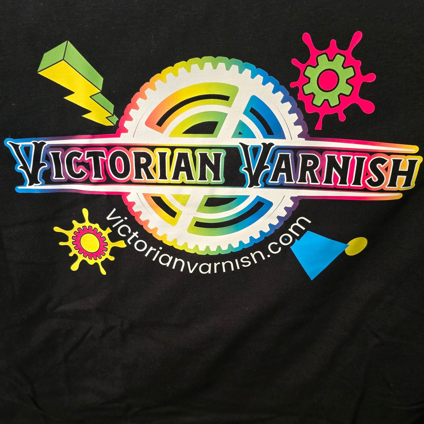 A close-up of a black garment featuring a colorful printed graphic with the text 'VICTORIAN VARNISH' and 'Victorianvarnish.com' over a rainbow gear design, alongside small bright decorative icons.