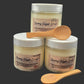 Three jars of Steamy Sugar Scrub with white lids and beige labels, arranged on a dark background with two wooden spoons.