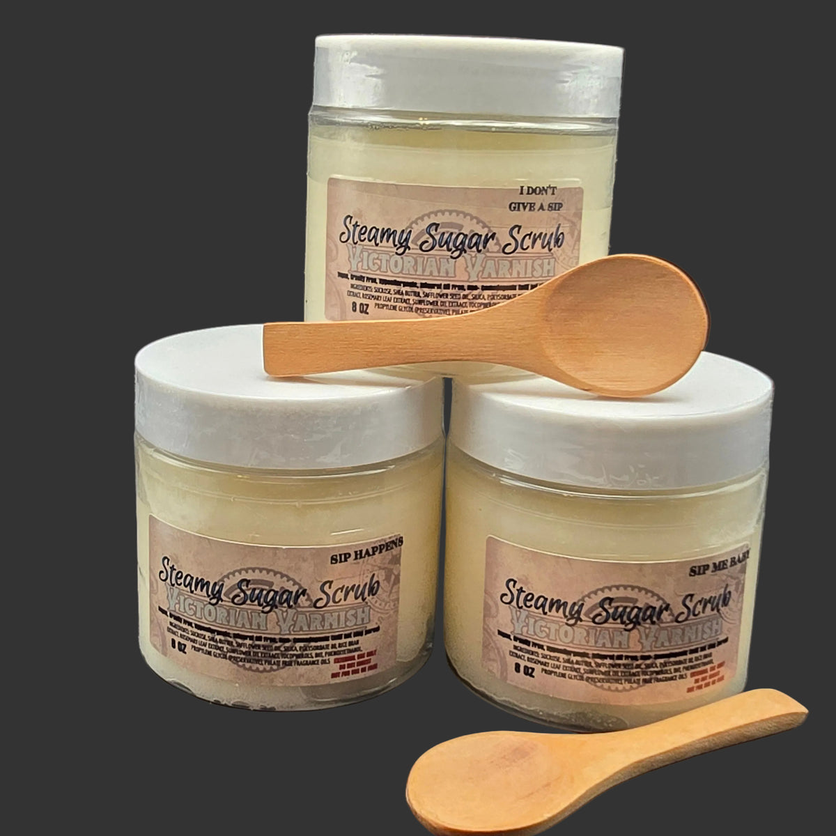 Three jars of Steamy Sugar Scrub with white lids and beige labels, arranged on a dark background with two wooden spoons.